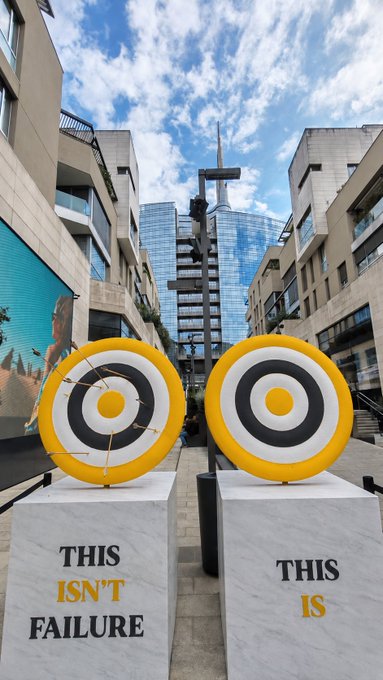 Two large yellow and black target sculptures stand on marble pedestals reading "THIS ISN'T FAILURE" and "THIS IS," visually reinforcing the post's message that every attempt—no matter how off-target—is worth pursuing. Set against a modern urban skyline, the artwork mirrors the resilience implied by the Binance account registration link, framing the pursuit of opportunity as something inherently valuable.