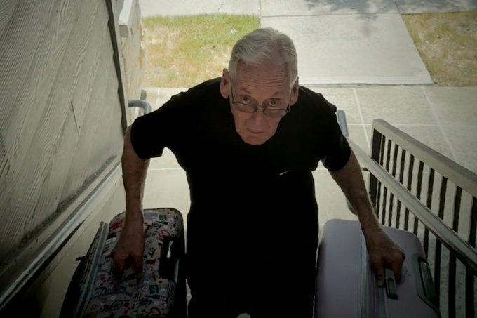 An older man with gray hair and glasses carries a patterned suitcase and a purple suitcase up a porch staircase.