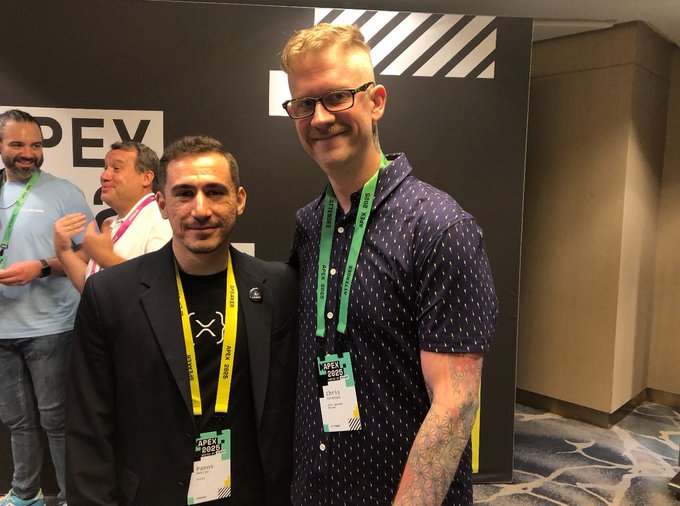 Two men pose together at what appears to be a convention, both wearing lanyards with 'APEX 2023' badges against a backdrop featuring the APEX logo.