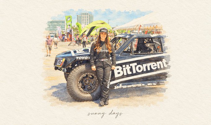 A woman poses confidently in a black BitTorrent-branded off-road racing truck, with "BitTorrent" prominently displayed on the side and "Toyo" tires visible, set against a sunny outdoor event backdrop captioned "sunny days." The image ties to the post "GM. Driven to connect." by showcasing BitTorrent's bold, adventure-driven branding that emphasizes connectivity and forward momentum.
