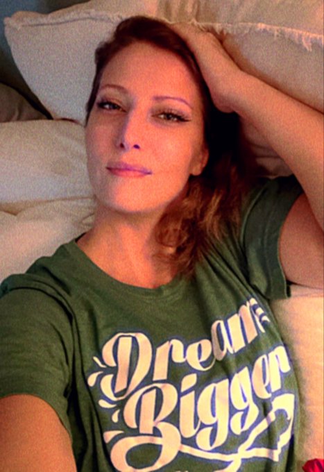 A woman with reddish-brown hair poses lying down against white pillows, wearing a green t-shirt with the text "Dream Bigg" printed in white retro-style lettering, matching the personal and cozy tone of her caption about the post not being algorithm-driven but rather a special request to her followers.