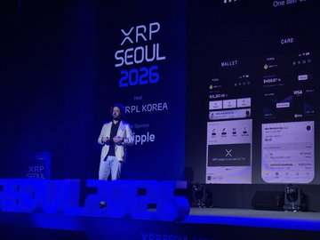 A speaker presents on stage at XRP Seoul 2026, with the "XRP SEOUL 2026" logo, "SPEAKER" text, and a Tria sponsor graphic displayed on the backdrop alongside mobile app mockups illustrating the neobank's "WALLET," "CARD," and "BORROW" features for XRP and RLUSD. The "SEOUL 2026" branding is also visible on large blue letters in the foreground, tying the keynote presentation about Tria's self-custodial finance app directly to the conference's setting.