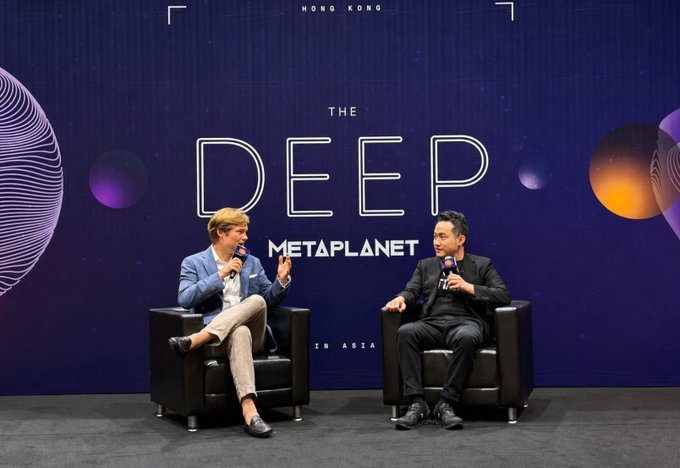 Two men seated in black chairs on a stage with a 'THE DEEP METAPLANET' backdrop, with 'HONG KONG' text at the top, appearing to be part of an interview panel at a Bitcoin Asia event.