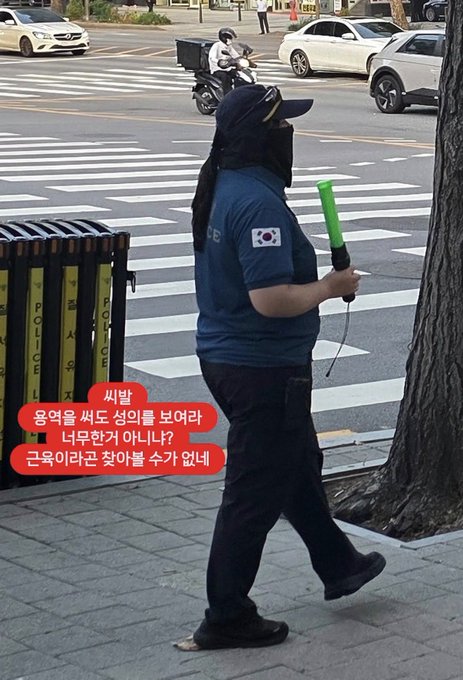 A man wearing a police uniform with a South Korean flag patch stands on a sidewalk holding a green traffic baton, with a Korean police barricade visible behind him. The image relates to the post's questioning of his appearance as a police officer, showing a stocky individual in official-looking attire near an urban crosswalk.