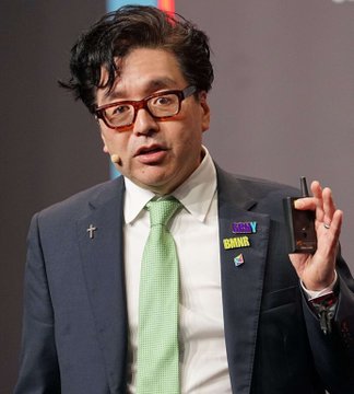 A photograph of a man in a dark blue suit, red glasses, and a green tie, holding a small black recording device or case.