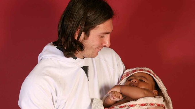 A man with long hair in a white hoodie smiles down at a newborn baby wrapped in a patterned blanket against a red backdrop.