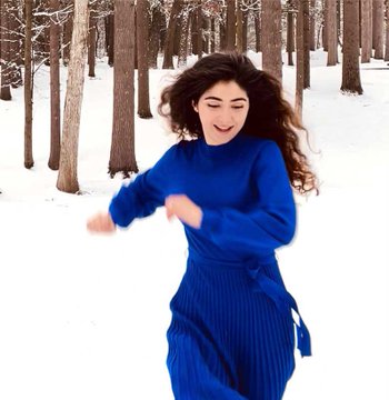 A woman with long dark hair twirling joyfully in a snowy forest, wearing a blue knit sweater and skirt.