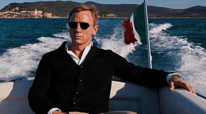 The image features a man wearing sunglasses, sitting inside a boat with his arm resting on the edge of the boat. He appears to be posing for a photo or enjoying the view from the boat. The background shows an ocean scene with waves, indicating that they are in a coastal area. There is also a flag visible in the background, which could possibly represent their location or the country where they are sailing.