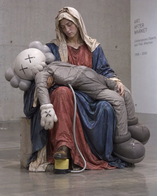 A contemporary art sculpture displayed at an 'ART AFTER MARKET: Contemporary Objects and Their Afterlives 1998-2005' exhibition, depicting a classical Madonna figure cradling a KAWS-style grey character with X-ed out eyes.