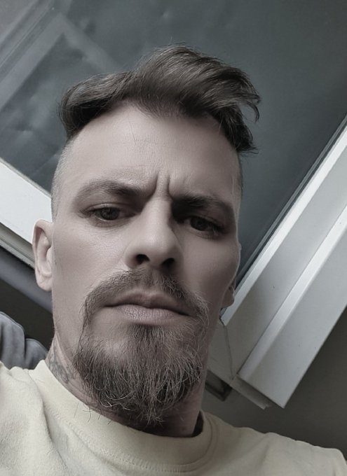 A black and white selfie of a man with styled hair, a goatee, and tattoos on his neck, looking serious at the camera.
