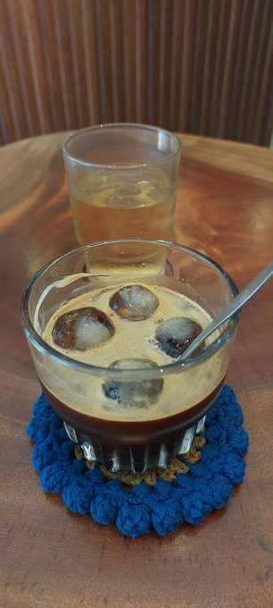 Two glasses of iced coffee or tea, with the front glass featuring large ice spheres, sit on a wooden table atop a blue crocheted coaster.