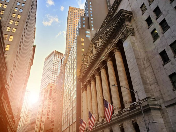 A low-angle street view of Wall Street's historic classical building with columns and American flags, bathed in warm sunlight from the left.