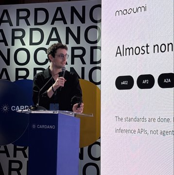 A man speaks at a podium labeled 'CARDANO' in front of a backdrop featuring the text 'CARDANO' and a screen displaying 'masumi' and 'Almost non'.
