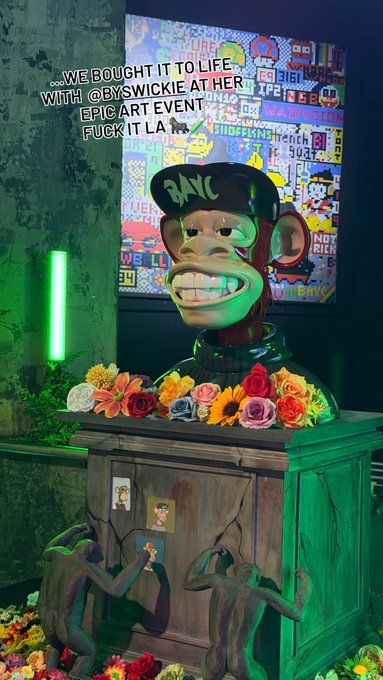 A sculpture of a BAYC (Bored Ape) head wearing a 'BAYC' cap, displayed on a wooden pedestal surrounded by colorful flowers and monkey figures, with overlay text reading '...WE BOUGHT IT TO LIFE WITH @BYSWICKIE AT HER EPIC ART EVENT FUCK IT LA.'