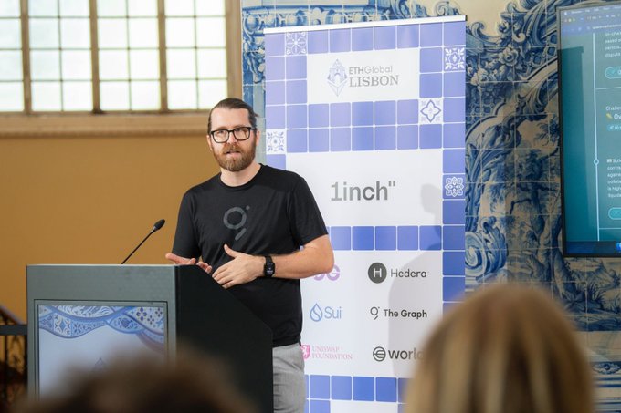 A bearded man with glasses speaking at a podium during the ETHGlobal LISBON event, with a banner displaying sponsors including Linch, Hedera, Sui, and The Graph.