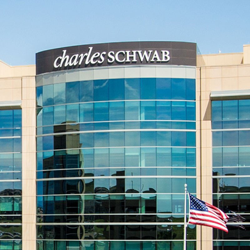 The top of a modern glass building displays the 'charles SCHWAB' logo, with an American flag flying in the foreground against a clear sky.