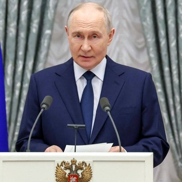 Vladimir Putin stands at a podium bearing the Russian coat of arms, wearing a dark suit and tie, delivering a speech in front of gray curtains.