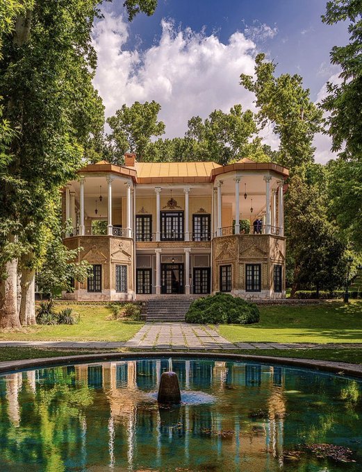 An elegant historic mansion or palace with arched windows and columns, painted in warm yellow and cream tones, reflected in a circular pool/fountain in the foreground, surrounded by lush green trees and a lawn.