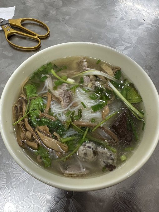 A bowl of Vietnamese noodle soup, likely pho, garnished with cilantro, green onions, and what appears to be mushrooms and pork, sitting on a table next to a pair of gold-handled scissors.