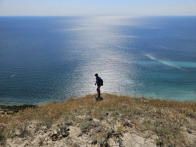 A hiker with a backpack stands on a grassy clifftop overlooking a vast, sunlit blue ocean.