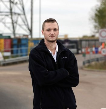 A man, presumably James Howells, stands with his arms crossed in front of what appears to be a waste management facility, wearing a black fleece jacket with "K.H.W" embroidered on it. The image relates to the accompanying post about Howells, the Welsh IT worker who famously discarded a hard drive containing thousands of Bitcoin.