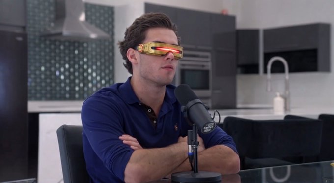 A man in a blue polo shirt sits in a modern kitchen with arms crossed, wearing gold and red Cyclops-style sunglasses, speaking into a "BAG CALLS" podcast microphone to announce a $500 SOL giveaway as a meme-style flex aimed at the timeline.