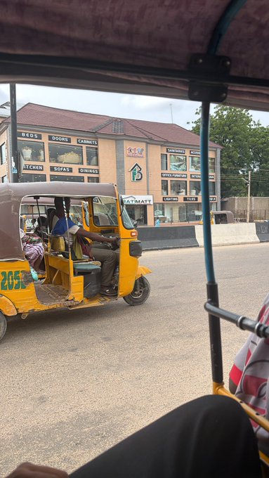 A yellow auto-rickshaw numbered "205" navigates a busy street past a "City Mart" building displaying signs for "BEDS," "DOORS," "CABINETS," "KITCHEN," "SOFAS," and "DININGS," capturing a candid moment of daily local transit. The scene reflects the everyday hustle of urban life, fitting the "day in the life" theme.