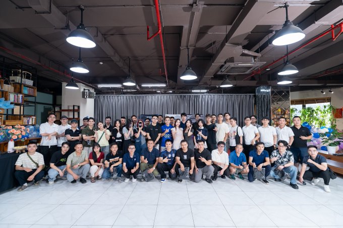 A large group photo of around forty people posing together in an industrial-style room with exposed ceiling pipes and hanging lights.