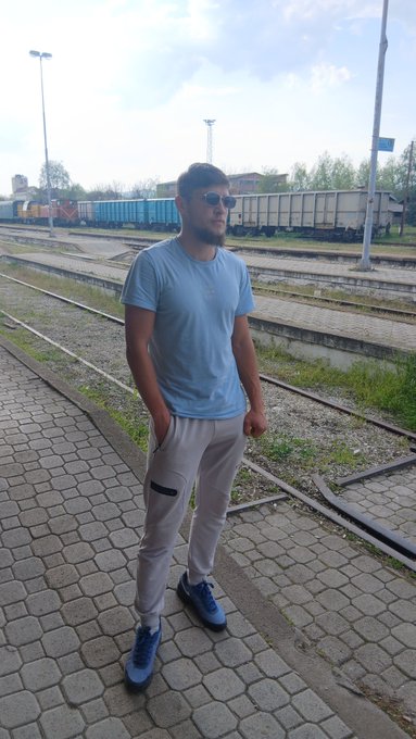 A man in a light blue t-shirt, gray pants, sunglasses, and blue sneakers stands casually on a train platform with his hand in his pocket, as freight train cars line the tracks behind him—a relaxed "Gm guys" pose perfect for a morning greeting.