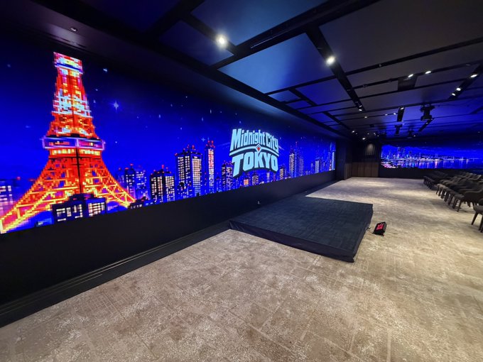 An interior view of a venue called 'Midnight City TOKYO' featuring a large LED wall displaying an illuminated Tokyo Tower and city skyline at night, with rows of seating and a stage area.