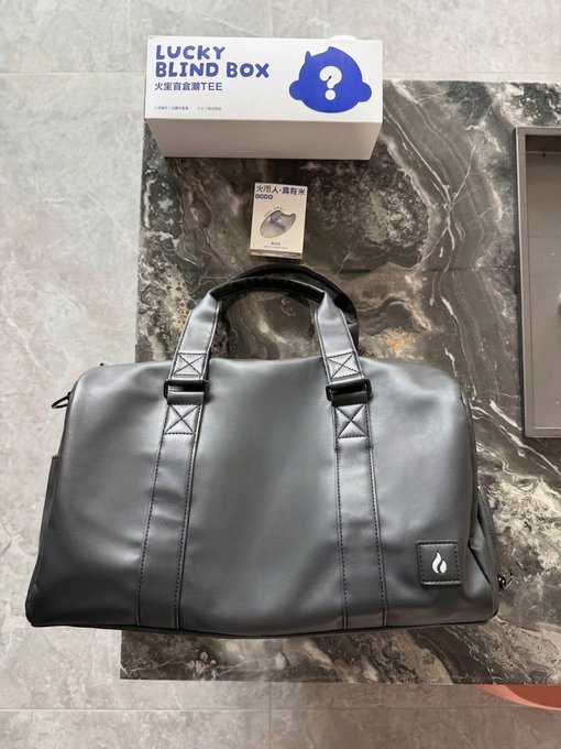 A black leather duffel bag with a small "Huobi" logo patch sits on a marble surface, accompanied by a "LUCKY BLIND BOX" promotional card featuring a blue mascot and a "400U" voucher, which appear to be Huobi's premium Mid-Autumn Festival gifts for users.