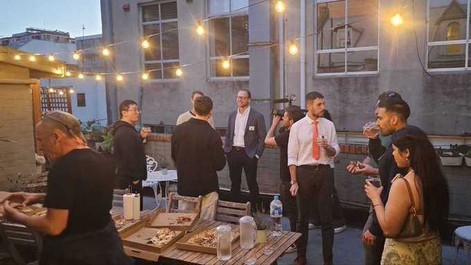 A photograph of a group of people socializing at an outdoor evening rooftop gathering with string lights, with pizza boxes visible on a table.