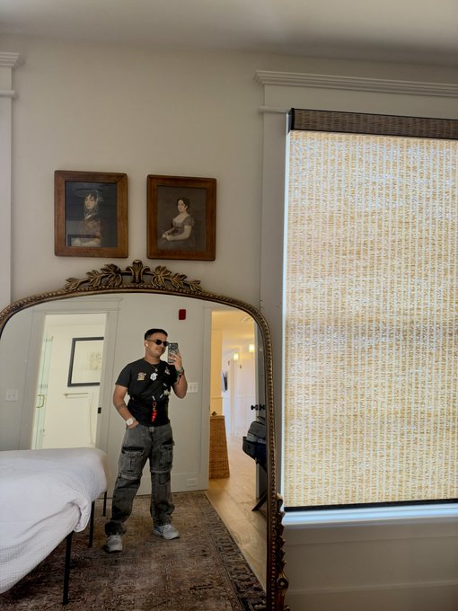 A man takes a mirror selfie in what appears to be a hotel room, wearing sunglasses, a black t-shirt, and gray cargo pants, with two classical portraits hanging above the mirror. The post's reference to "Beeple Studios" suggests this is likely at the artist Mike "Beeple" Winkelmann's residency space, where such portrait-adorned, artist-curated rooms are part of the experience.