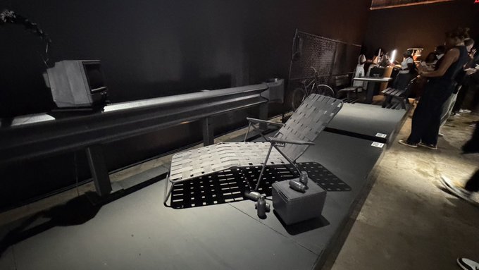 A metal chair and small weight rest on a platform at what appears to be a Jay-Z themed pop-up exhibit, with a few visitors visible in the background. The industrial, minimalist setup reflects the "Chillin in the #JAYZ pop-up in Dumbo" experience, evoking a virtual, metaverse-like atmosphere reminiscent of the @OthersideMeta space.