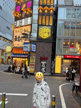 A person wearing a black and white patterned jacket and an emoji cap standing on a street in front of various Japanese storefronts with neon signs.