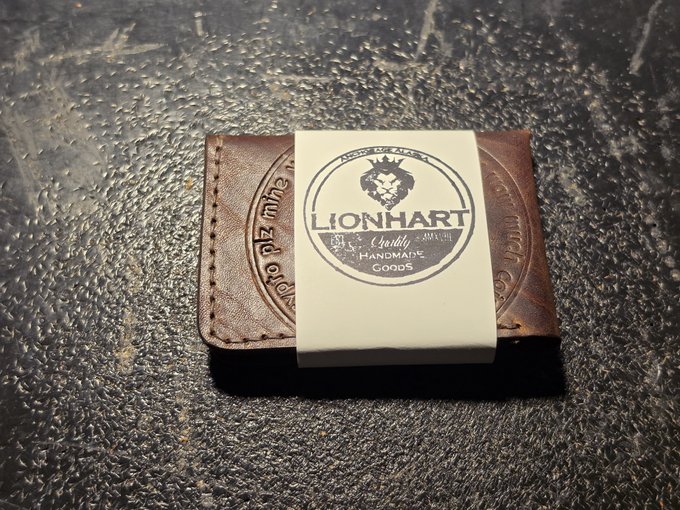 A brown leather bifold wallet with a 'LIONHART Quality Handmade Goods' branded paper band wrapped around it, sitting on a dark textured surface.