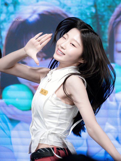 A young woman with long dark hair smiling and posing with her hand near her face, wearing a white sleeveless top and red pants, with 'SoCute!' written in the top left.