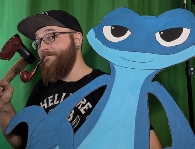 A bearded man wearing glasses and a cap poses with a large painted cutout of a blue cartoon character, holding a prop axe in front of a green screen.