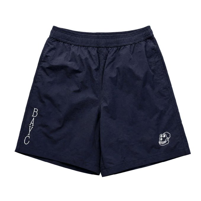A pair of plain navy blue nylon shorts is displayed against a white background, featuring the word 'BAYC' written vertically in white on the left leg and a small white cartoon graphic on the right leg.