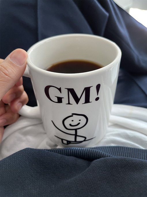 A hand holds a white mug filled with dark coffee, printed with 'GM!' and a stick figure in a meditative yoga pose.