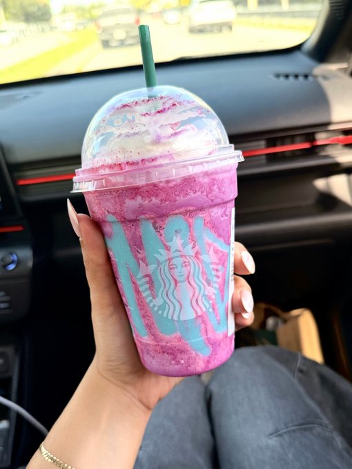 A hand holding a purple and blue Starbucks 'Unicorn' Frappuccino with whipped cream and a green straw inside a car.