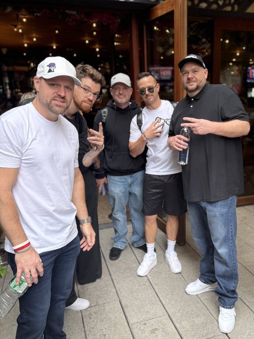 Five men pose together outdoors, with two making hand gestures; one wears a white cap, another holds a phone, and one has a water bottle.