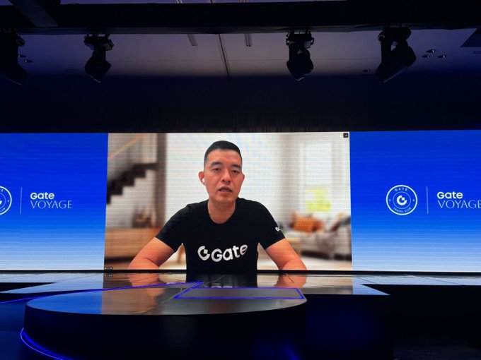 A man wearing a black 'Gate' t-shirt appears on a large studio screen during a 'Gate VOYAGE' event.
