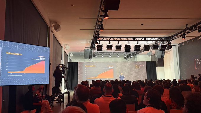 A view from the audience of a large conference hall where a presenter is speaking, with screens displaying a "Tokenization" growth chart rising to $45.1B.