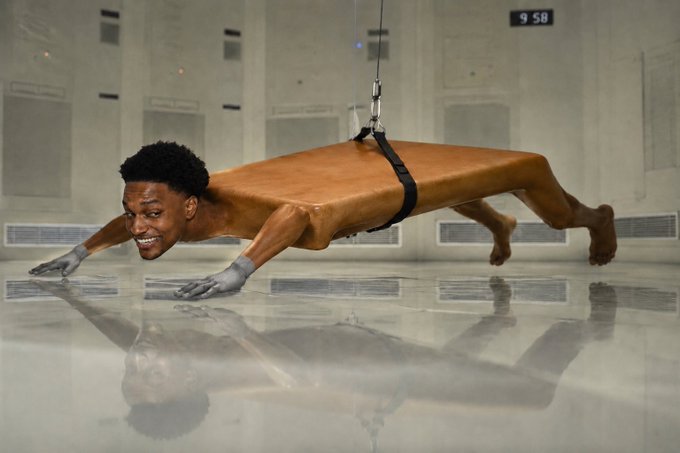A surreal, edited image shows a person's body in a push-up position wearing only a harness, with their head replaced by a smiling man's face, appearing to float or be suspended above a reflective floor.