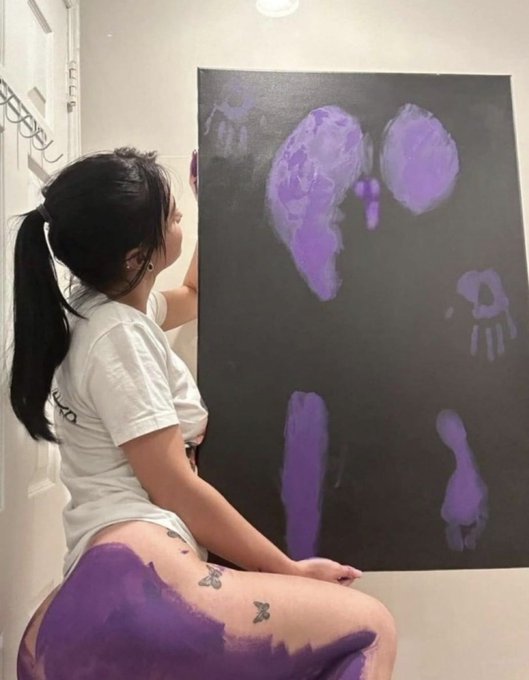 A woman with a ponytail in a white shirt sits facing a black canvas with purple handprint-like paint smears, with purple paint also visible on her leg.