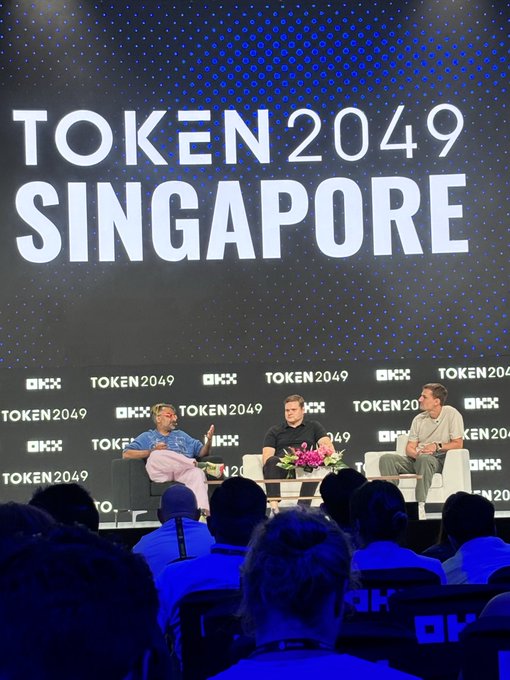 Three people sit on stage at the TOKEN 2049 Singapore conference, with the event name displayed on a large screen behind them.