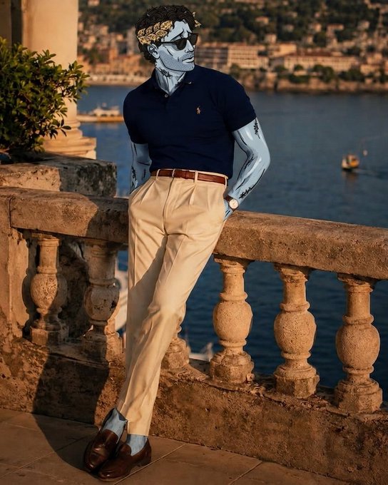 A fashion model with a classical stone bust painted on his skin, wearing sunglasses, a navy polo, and khaki pants, leaning against a stone balustrade by the sea.