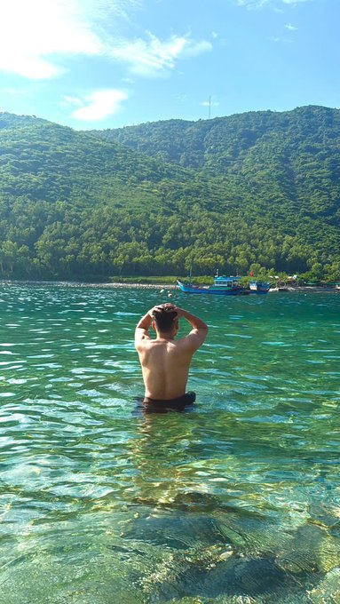 A photograph of a shirtless man standing waist-deep in clear, turquoise ocean water with his hands on his head, facing a lush green mountainous coastline with a couple of small boats nearby.