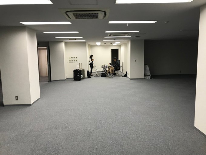 Two people sit in folding chairs at a small table in an otherwise empty, carpeted office space with luggage nearby, suggesting a modest, early-day setup. The sparse surroundings contrast with the post's theme of humble beginnings for the world's largest bitcoin exchange.
